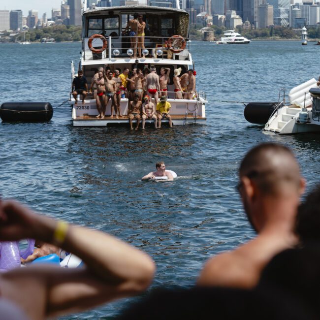 A group of people on a boat party in a harbor, with boats nearby and a city skyline in the background. A person is swimming in the water, surrounded by onlookers on nearby boats.