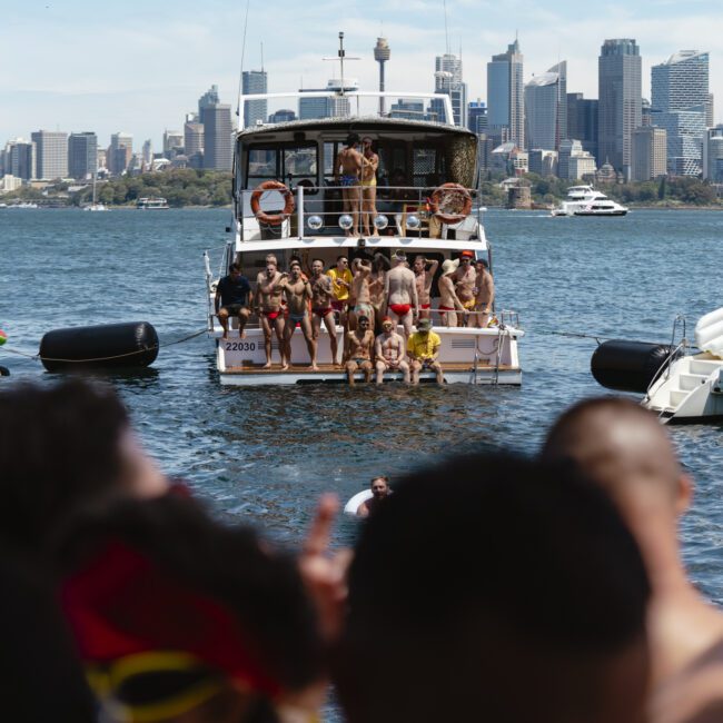 A group of people in swimwear enjoy a party on a boat in a harbor with a city skyline in the background. Other boats with more partygoers surround the main party boat. The scene is festive and vibrant with clear skies above.