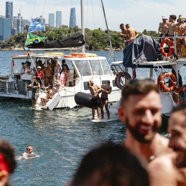 People enjoying a lively boat party on a sunny day. Two boats are filled with people in swimwear, some in the water, with a city skyline and greenery in the background. Bright and festive atmosphere.