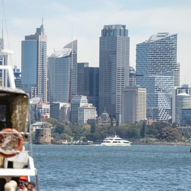 A city skyline featuring a mix of modern skyscrapers and older buildings. In the foreground, there is a body of water with a boat and a small lighthouse. Trees are visible along the shoreline. The sky is partly cloudy.