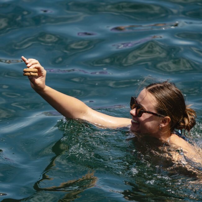 A person with sunglasses is swimming in clear blue water, smiling and holding up a brown object, possibly food, in one hand. The person appears to be enjoying the sunny day.