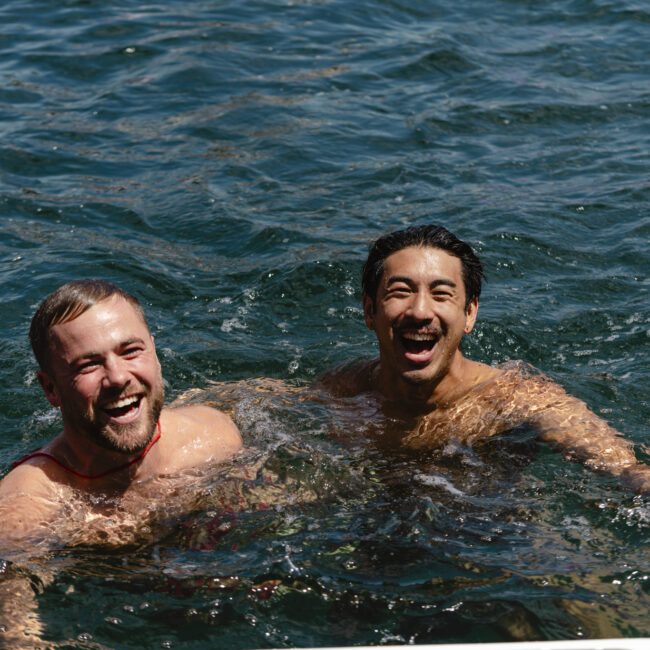 Two men are smiling and swimming in a body of water under bright sunlight. The water is dark blue and rippled. Both appear to be enjoying the moment.