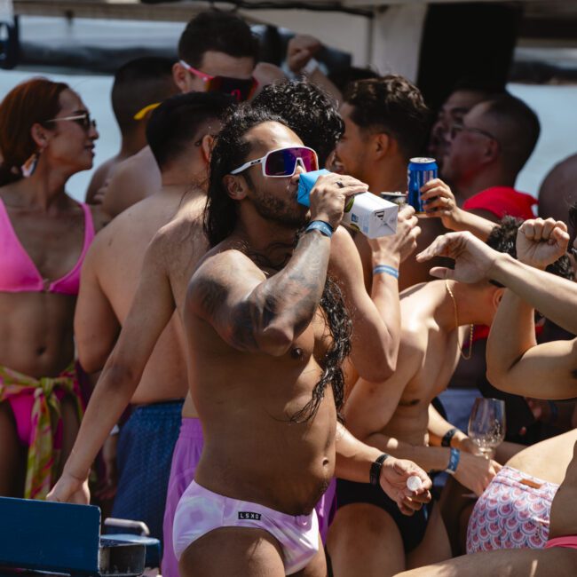 A lively group of people in swimwear enjoying a party on a boat. Some are holding drinks, others are dancing, and most are wearing sunglasses. The atmosphere is festive and colorful, with sunlight illuminating the scene.