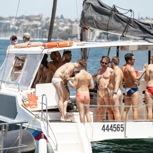 A group of people in swimsuits are gathered on a boat, socializing and holding drinks. The boat is on a body of water, with a cityscape visible in the background. Bright and sunny day.