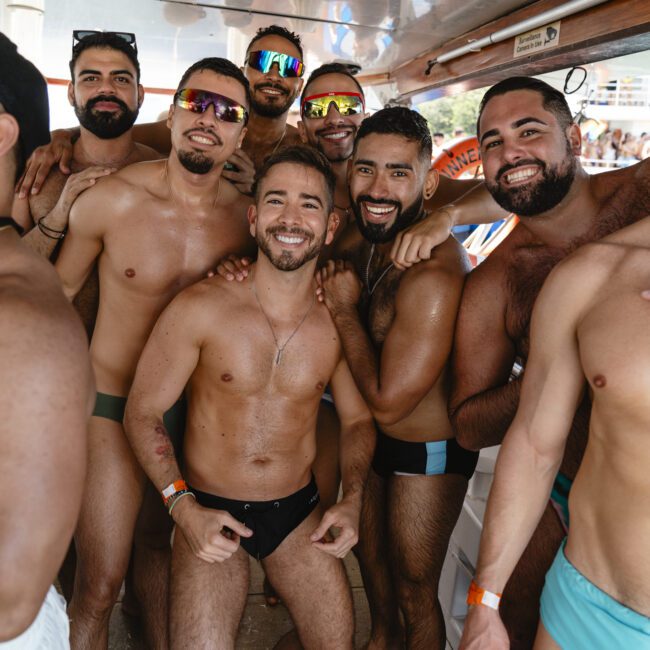 A group of eight men smiling and posing together on a boat. They are wearing swim trunks and sunglasses. There is festive decor and other people visible in the background.