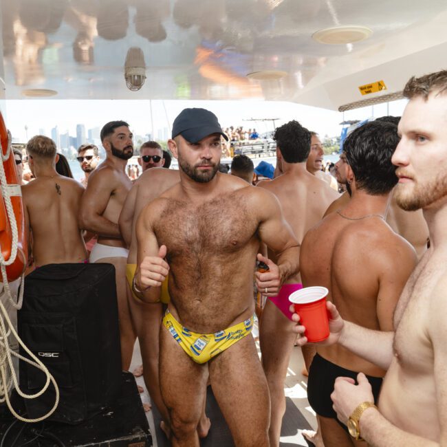 A group of shirtless men are gathered on a boat. One man in the center is wearing a hat and yellow swimsuit, giving a thumbs up. Another is holding a red cup. The boat is crowded, and the city skyline is visible in the background.