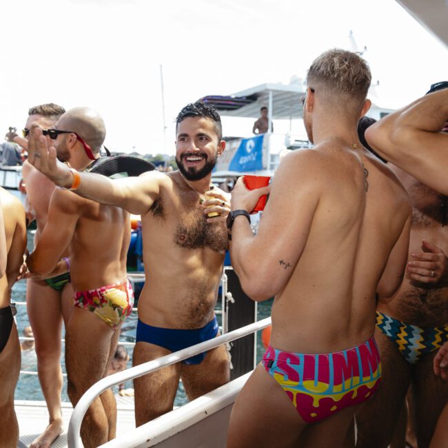 A group of men wearing colorful swimwear are on a boat, smiling and chatting. The atmosphere is lively and summery, with clear skies in the background. Some men hold drinks, enjoying the social gathering.