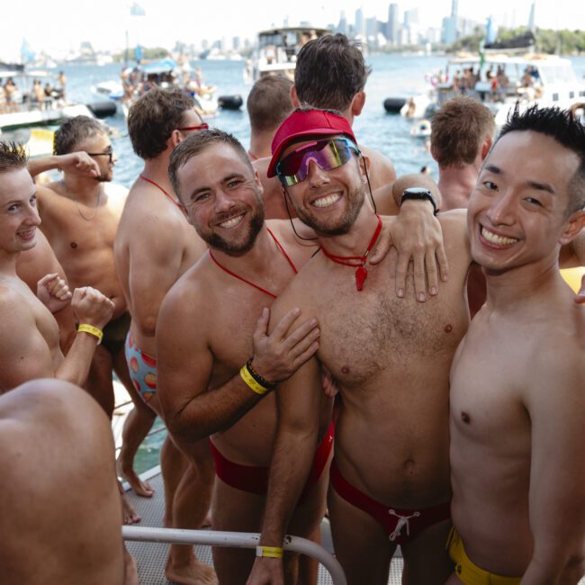 A group of people in swimsuits are smiling and posing on a boat surrounded by water and other boats. It's a sunny day and the city skyline is visible in the background.