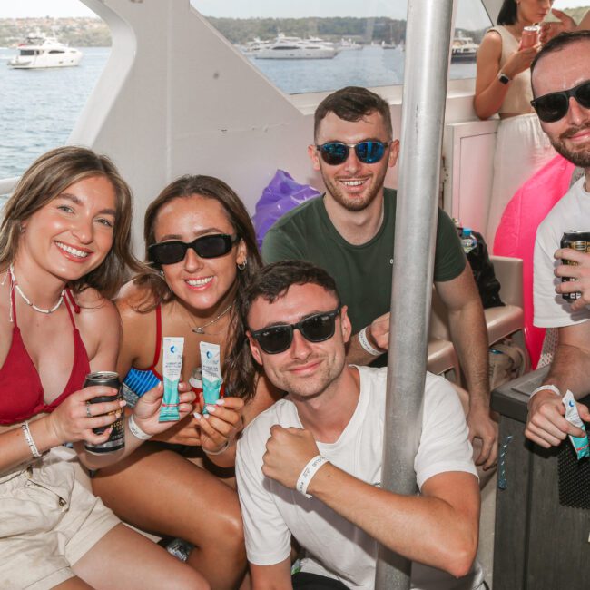 A group of five people wearing sunglasses pose together on a boat. Three are seated, two standing behind them, each holding a can. The background shows water and several boats under a clear sky. Everyone is smiling.