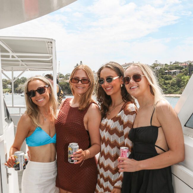 Four women in summer attire stand on a boat deck holding drinks, smiling at the camera. They're wearing sunglasses and enjoying a sunny day. The backdrop includes water and a hilly landscape under a partly cloudy sky.