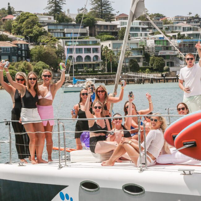 A group of people enjoys a sunny day on a boat, holding drinks and posing for the camera. They are surrounded by water with houses and trees in the background. Some are standing while others are seated, creating a lively and cheerful atmosphere.