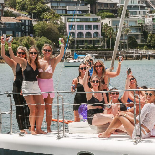 A group of women in swimsuits and summer attire are smiling and raising their drinks on a boat. They are enjoying a sunny day on the water, with houses and trees visible in the background.