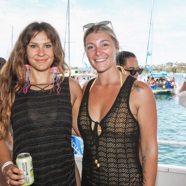 Two women smiling at an outdoor event on a boat. They are wearing black sleeveless tops and holding drinks. The background features people and boats in the water, with sunny weather.