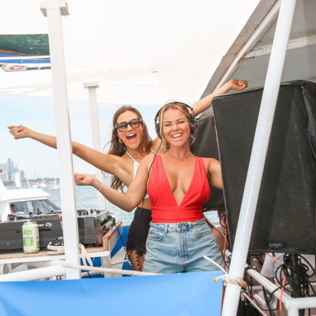 Two women on a boat are enjoying a sunny day. One is wearing sunglasses and a black outfit, and the other is in a red top with headphones. They are smiling and appear to be having fun near DJ equipment, with water and cityscape in the background.