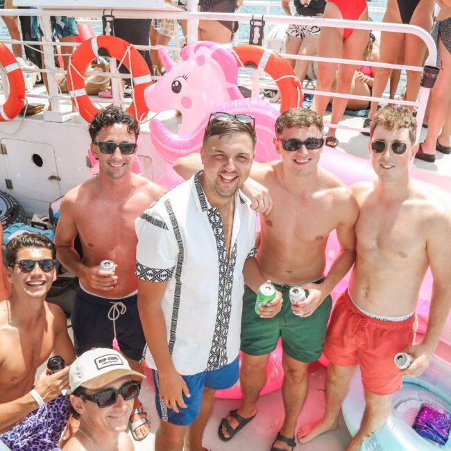 A group of friends on a boat deck, enjoying a party. They are wearing swimwear and sunglasses, holding drinks. Pink inflatable floats, including a flamingo, are in the background. The sun is shining, and the atmosphere is festive.