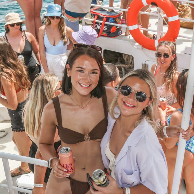 Two women smiling and holding drinks on a boat. One is wearing a brown bikini, and the other is in a light blue shirt and sunglasses. Behind them, a group of people enjoy a sunny day on the water, with a life preserver visible.