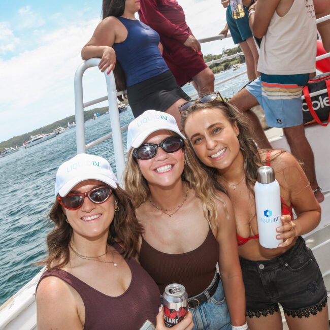 Three smiling people wearing matching white hats and sunglasses stand on a boat. They hold drinks and enjoy a sunny day on the water. Other passengers and boats are visible in the background.