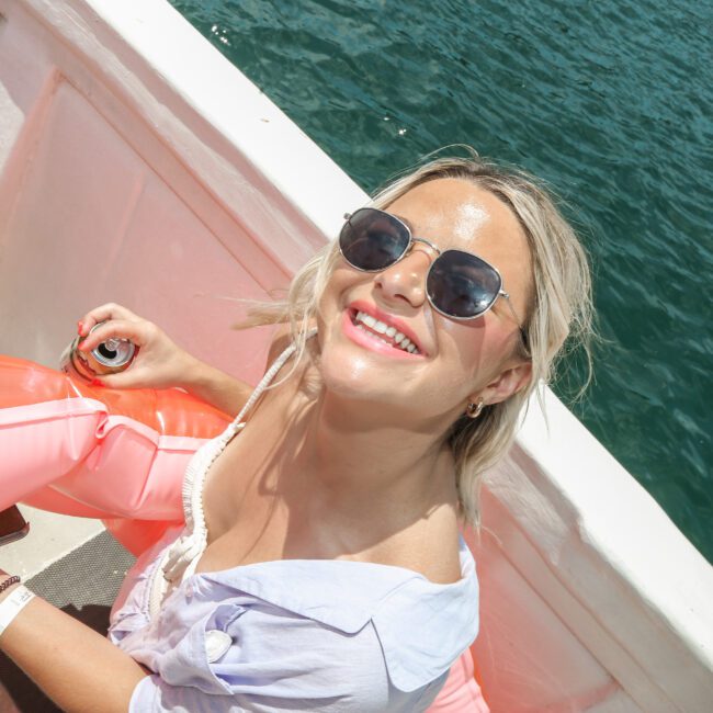 A woman with blonde hair and sunglasses is smiling and sitting on a boat. She is holding a pink inflatable object and has a can in her hand. The background shows blue water.