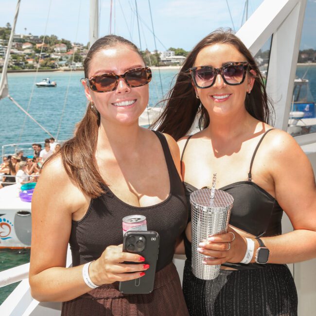 Two women in sunglasses smile while holding drinks on a boat deck. They wear black dresses and pose in front of a scenic waterfront with another boat visible in the background. The scene suggests a sunny, leisurely day on the water.