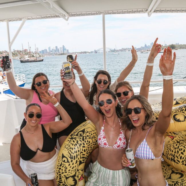 A group of six women in swimwear, wearing sunglasses, are celebrating on a boat. They are surrounded by inflatable rings and are holding drinks. The background features a waterfront view with city buildings and a clear sky.