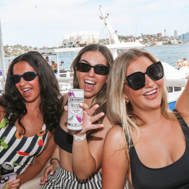 Three women in sunglasses, smiling and holding drinks, pose for a photo on a boat. The background features clear skies, a coastline, and a body of water.