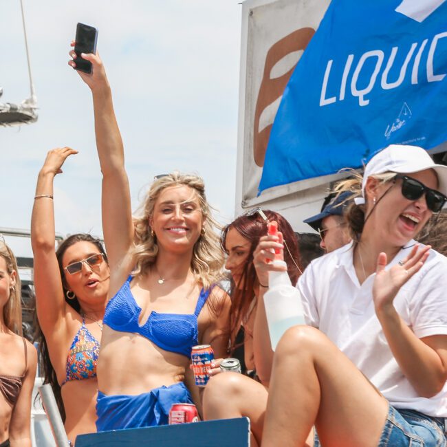 A group of people smiling and waving on a boat under a blue sky. They are wearing swimsuits and sunglasses, enjoying the outdoors. A blue "Liquiv" banner is partially visible in the background.