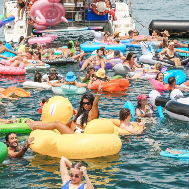 A lively scene of a crowd enjoying a boat party, floating on colorful inflatables in the water. People are interacting and relaxing under the sun, with inflatable shapes including a duck, rings, and a pink flamingo. A boat is docked in the background.