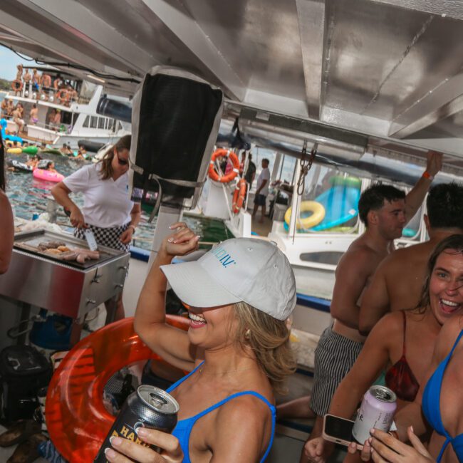 A lively group of people dance and enjoy drinks on a boat. Some wear swimsuits and hats, smiling and laughing. The background has boats on the water with more people visible. The scene is festive and captures a fun, summer atmosphere.