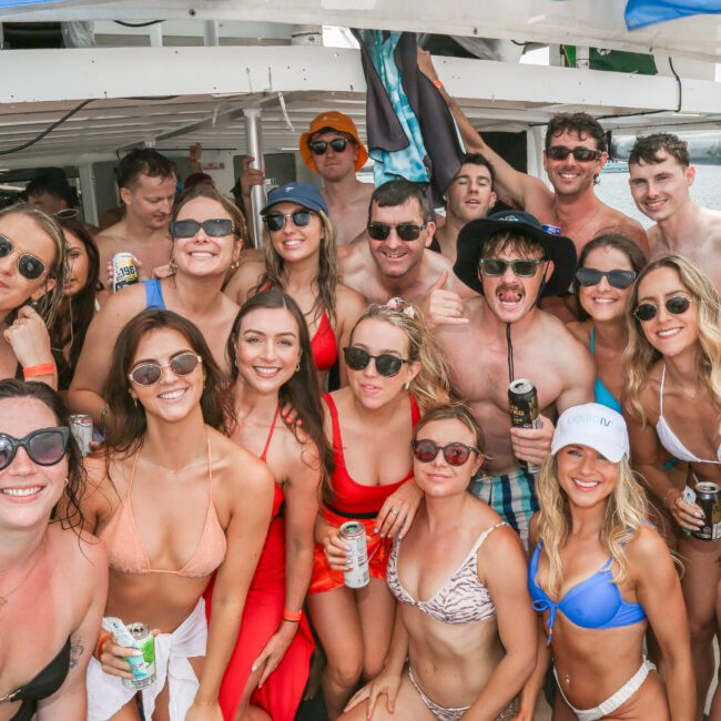 A large group of people dressed in swimwear is gathered on a boat, smiling and holding drinks. They appear to be enjoying a sunny day on the water, with some wearing hats and sunglasses.