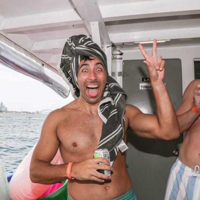 A man with a towel wrapped around his head excitedly holds up two fingers and a drink can, standing on a boat. Another man beside him is laughing with his eyes closed. The city skyline is visible in the background over the water.