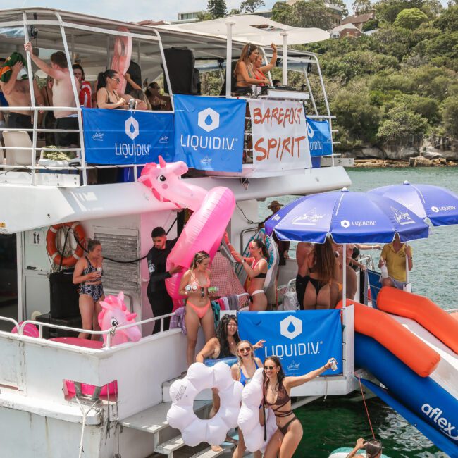 A lively boat party with people in swimwear, surrounded by inflatables and blue Liquidiv banners. Attendees enjoy music, dancing, and water slides, with some people on a paddleboard in the water.