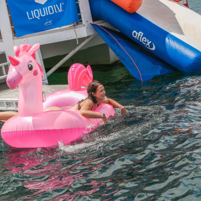 Person smiling on a pink unicorn float in the water, with a boat and waterslide in the background.