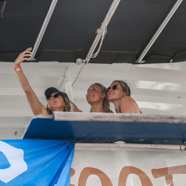 Three individuals on a balcony take a selfie together. They are smiling and enjoying the moment. A blue and white banner with a logo is partially visible below them.