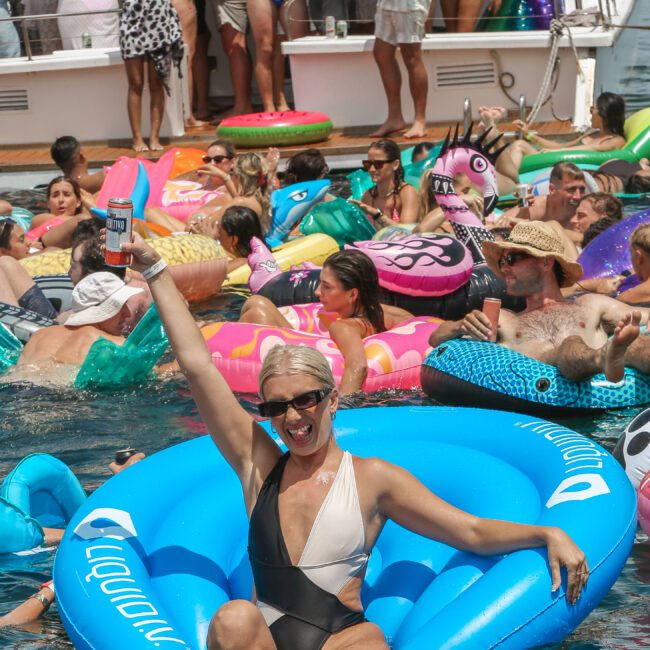 A person in a black and white swimsuit lounges on a blue inflatable chair in a pool, holding up a canned drink. The background is filled with people on inflatables, socializing near a boat. It's a lively and festive atmosphere.