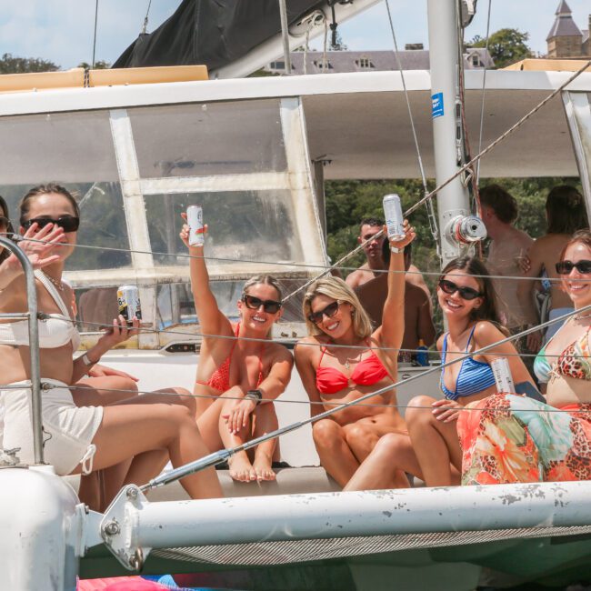 A group of women in swimwear sit and stand on the deck of a boat, smiling and waving at the camera. They are holding drinks and enjoying the sunny day. The boat is anchored near a shoreline with trees and buildings visible in the background.