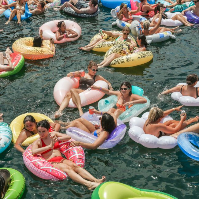 A group of people relaxing on colorful inflatable floaties in a body of water. They are enjoying the sun, with some wearing sunglasses and swimwear. The floaties are in various shapes and colors, creating a vibrant and lively scene.