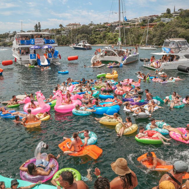 A vibrant scene of a crowded floating party, with numerous people enjoying themselves on colorful inflatable rafts and floats in the water. Several boats are anchored nearby, and the shoreline is lined with greenery under a clear sky.