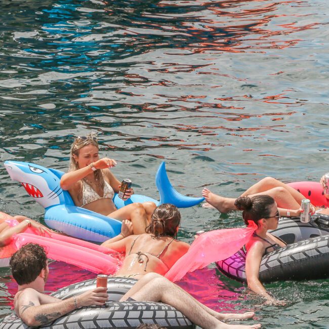 A group of people relaxing on colorful inflatables in the water. Some hold drinks while lounging on pink and gray tubes, and one inflatable resembles a shark. The scene conveys a fun, leisurely gathering.