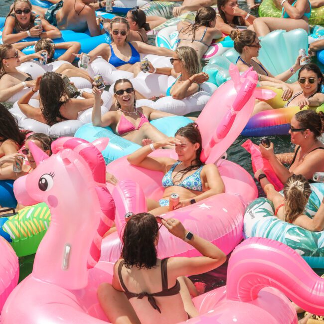 A crowded group of people relaxing on inflatable pool floats in the water. The floats are colorful and shaped like various animals, including unicorns and flamingos. The sun is shining, and everyone appears to be having a good time.