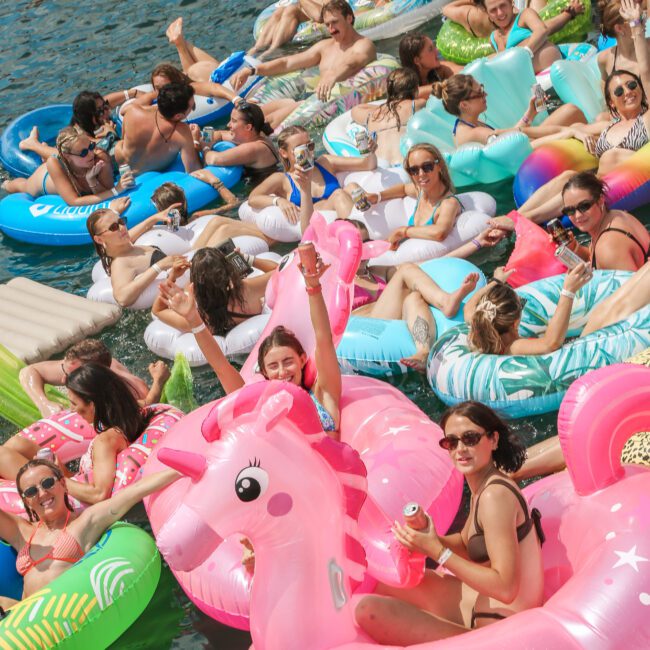 A crowd of people relax on colorful inflatable pool floats in a body of water. They are sunbathing, smiling, and enjoying the scene. Notable floats include a large pink unicorn and various vibrant designs.