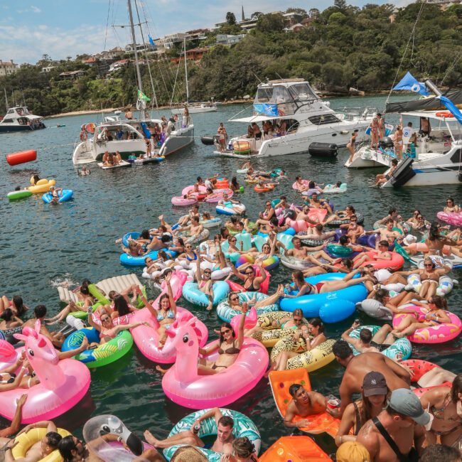 A large crowd of people relaxing on colorful inflatable floaties in the water, surrounded by motorboats. The scene is lively, with many bright inflatables, including flamingos and pool rings. Trees and houses are visible in the background.