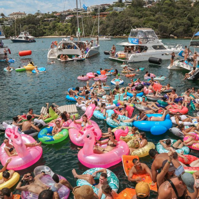 A large group of people enjoying a party on a lake, floating on colorful inflatable rafts, including flamingos and pizza slices. Several boats are anchored nearby, with people socializing and relaxing under a sunny sky.