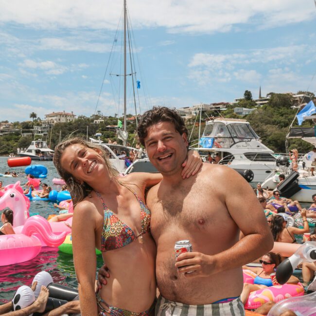 A smiling man and woman in swimwear stand on a boat, with numerous inflatable floaties and people in the water around them. The scene includes several anchored boats, a clear blue sky, and coastal greenery in the background.