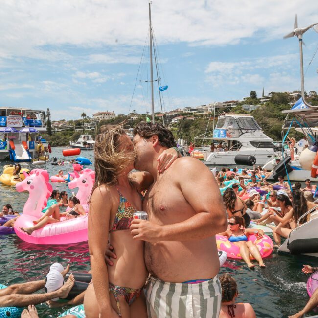 A couple kisses on a yacht surrounded by people on inflatable floats in the water. Various boats and yachts are anchored nearby. The scene is crowded and festive, with colorful inflatables like pink flamingos adding a playful vibe.