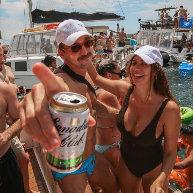 A group of people are enjoying a boat party on the water. Two people in the foreground, wearing swimwear and caps, are holding drinks. The scene is lively, with more people chatting and swimming nearby, surrounded by yachts.