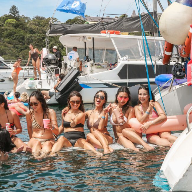 A group of six women in swimsuits sit at the edge of a boat, enjoying drinks and smiling. They are surrounded by inflatables and other boats on a sunny day. The water is clear, and trees are visible in the background.