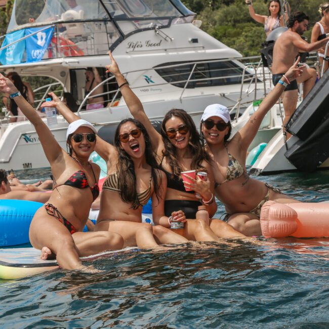 Four people in swimwear are sitting on a floating mat in the water, posing cheerfully with arms raised. They're surrounded by inflatable pool toys, with boats visible in the background. The scene is sunny and festive.