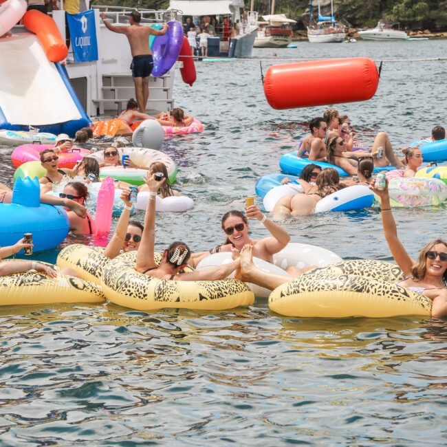 A group of people enjoying a sunny day in a scenic harbor area, floating on colorful inflatable tubes and loungers. They are smiling and holding drinks. Boating and lush greenery are visible in the background.