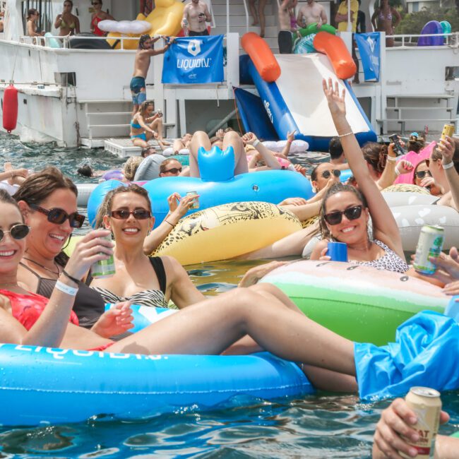 A group of people in swimsuits are relaxing on inflatable floats in the water, holding drinks and smiling at the camera. A large boat is in the background, with a slide extending into the water. Trees are visible in the distance.