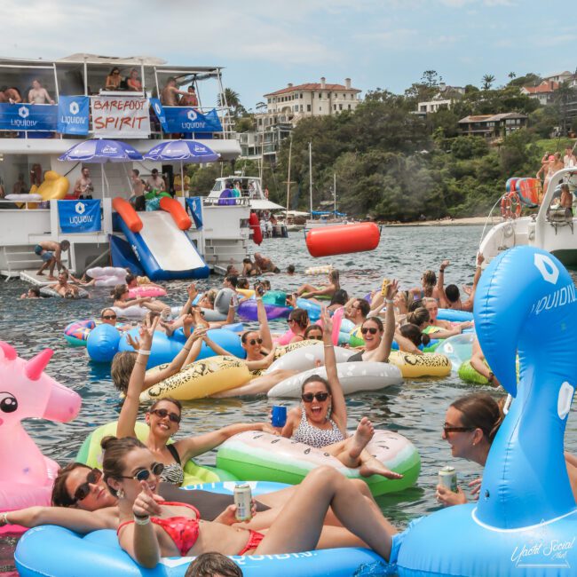 A lively group of people relax on colorful inflatable pool floats in the water near a boat with "Barefoot Spirit" signage. Many are smiling and holding drinks. The scene is festive, with a backdrop of other boats and a scenic shoreline.